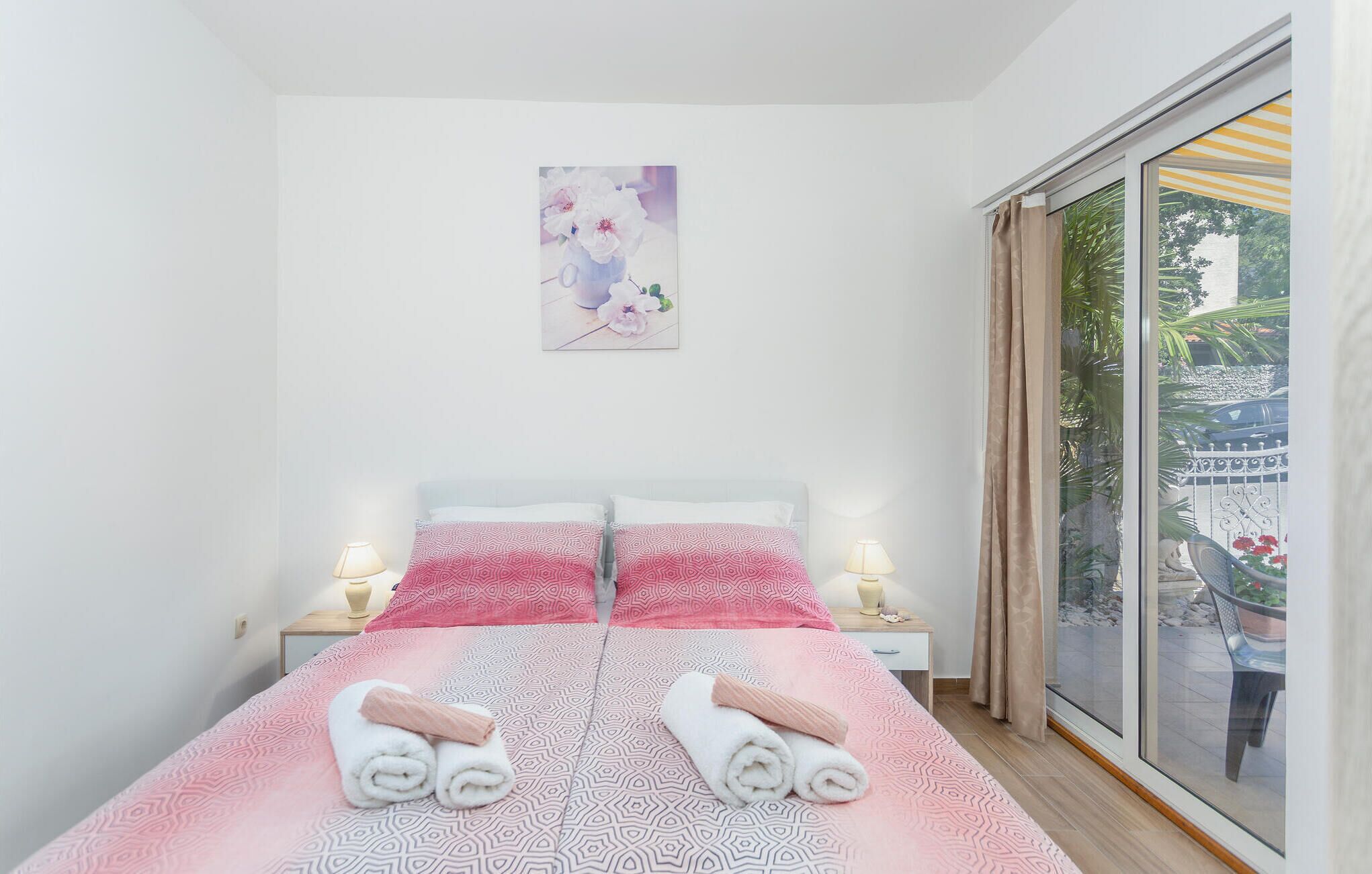 A bedroom with a bed, two bedside lamps, a window with a view of greenery, and a painting on the wall.