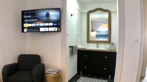 A modern living room with a flat-screen TV displaying a movie, a black armchair, a small round side table with books, and a black and white patterned rug leading to a bathroom with a vanity and mirror. - Perrosian villa with Jacuzzi, Sauna, Hammam, Home Cinema (Perros-Guirec)