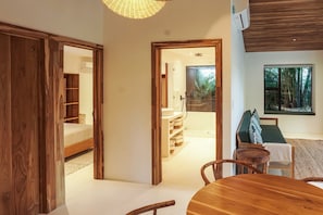 A spacious room with a wooden dining table, a sofa with cushions, and a view of a tropical landscape through a large window.