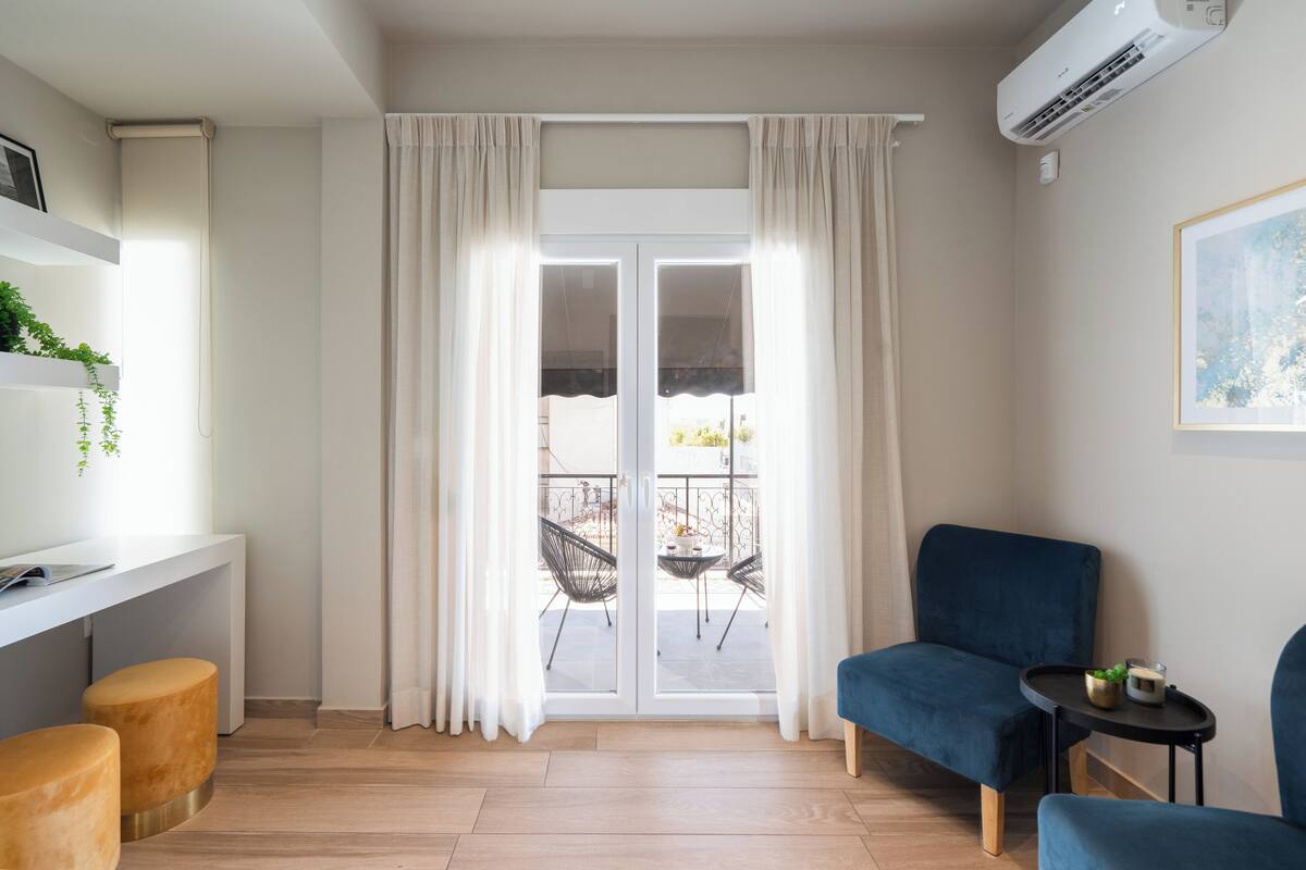 Ein modernes Zimmer mit einer Schiebeglasdoor zu einem Balkon, einem weißen Schreibtisch mit Stuhl, einem blauen Sofa und einem kleinen Tisch mit Pflanze.
