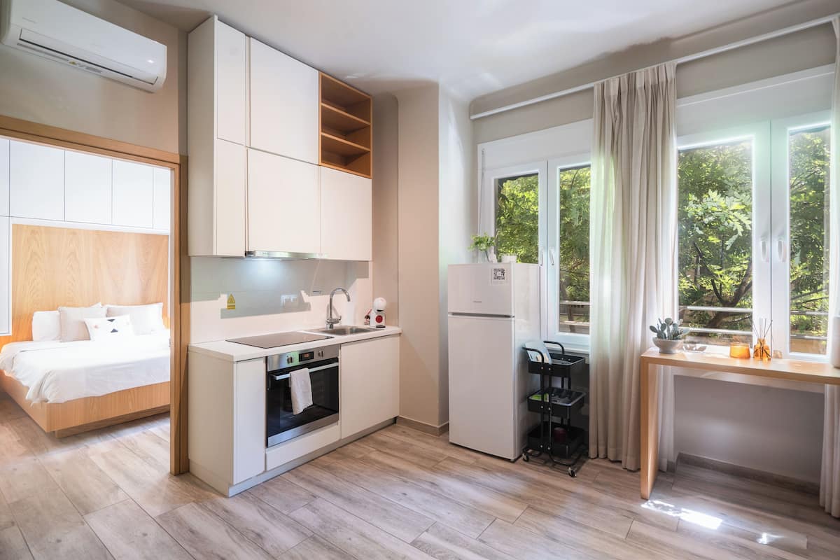 Ein kompaktes Hotelzimmer mit Küchenzeile, einem Bett mit Holz Kopfteil, einem Fenster mit Vorhängen und Blick auf Grünfläche.