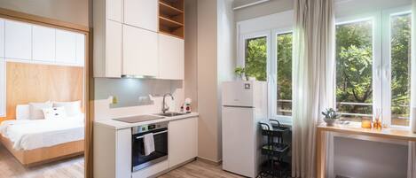 Une chambre d’hôtel compacte dotée d’une kitchenette, d’un lit avec une tête de lit en bois, d’une fenêtre avec des rideaux et d’une vue sur la verdure environnante.