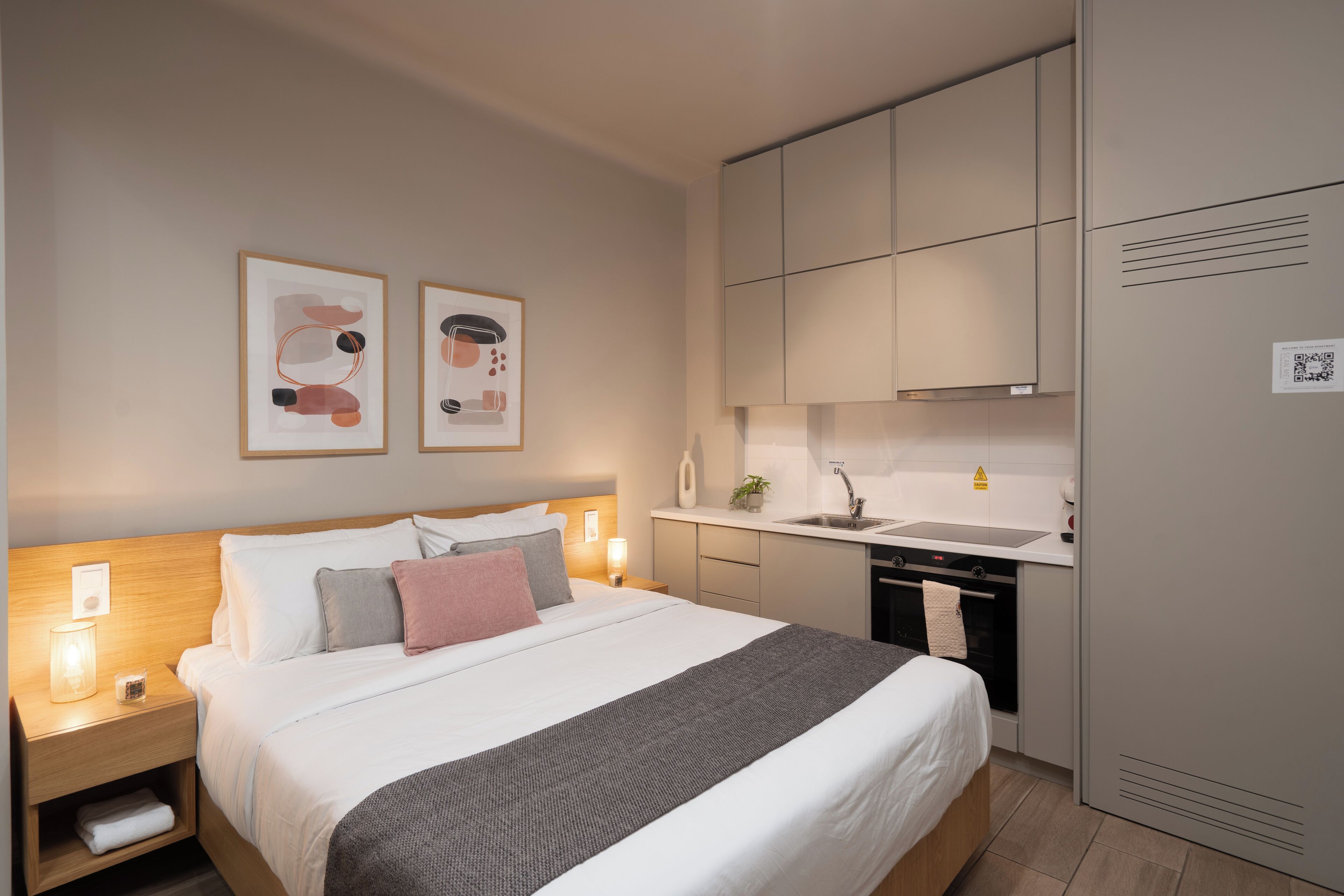 A hotel room with a bed, bedside tables, a nightstand, a small shelf, a wall-mounted shelf, a bedside lamp, a framed picture, a kitchen area with a sink, a stove, and cabinets.