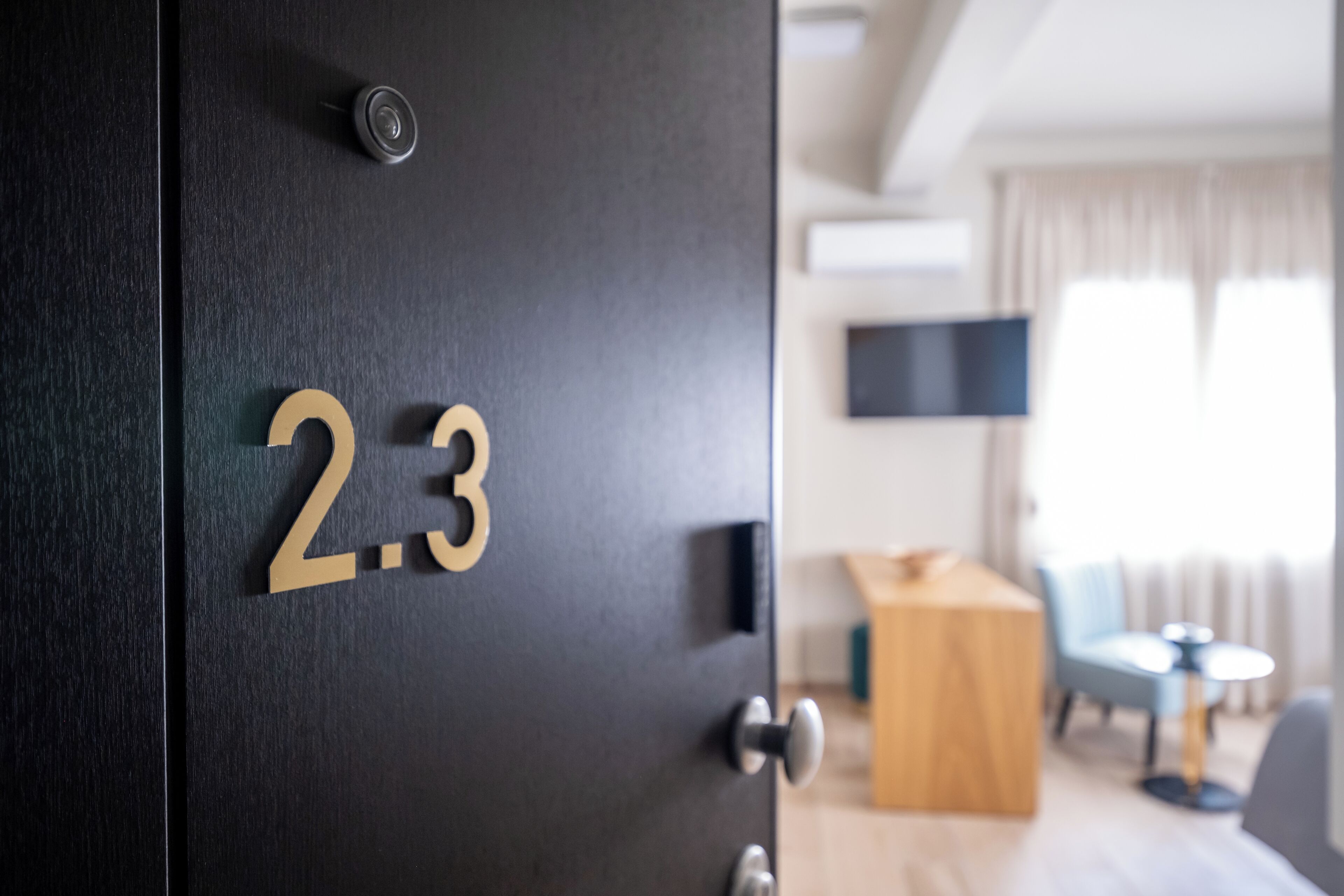 A hotel room door with the number 23, a modern interior with a flat-screen TV, a wooden table, and a blue chair.