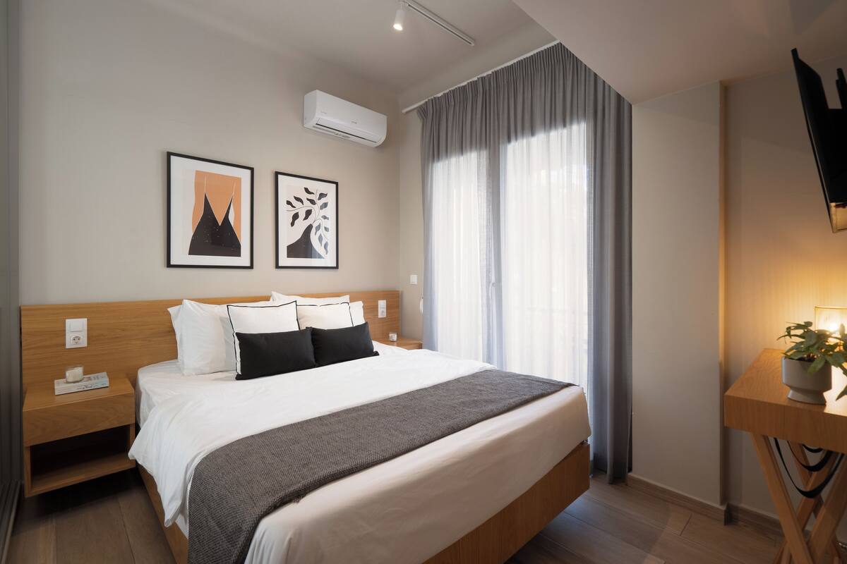 Ein modernes Hotelzimmer mit einem großen Bett, einem Nachttisch aus Holz, einer Stehlampe, einer Topfpflanze und einer Wandklimaanlage.