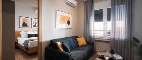 Habitación de hotel moderna con sofá negro, una mesita con una revista, planta en maceta y una cama con cabecero de madera.