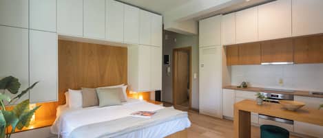 A modern bedroom with a large bed, bedside tables, a bedside plant, and a view into the adjacent kitchen area.