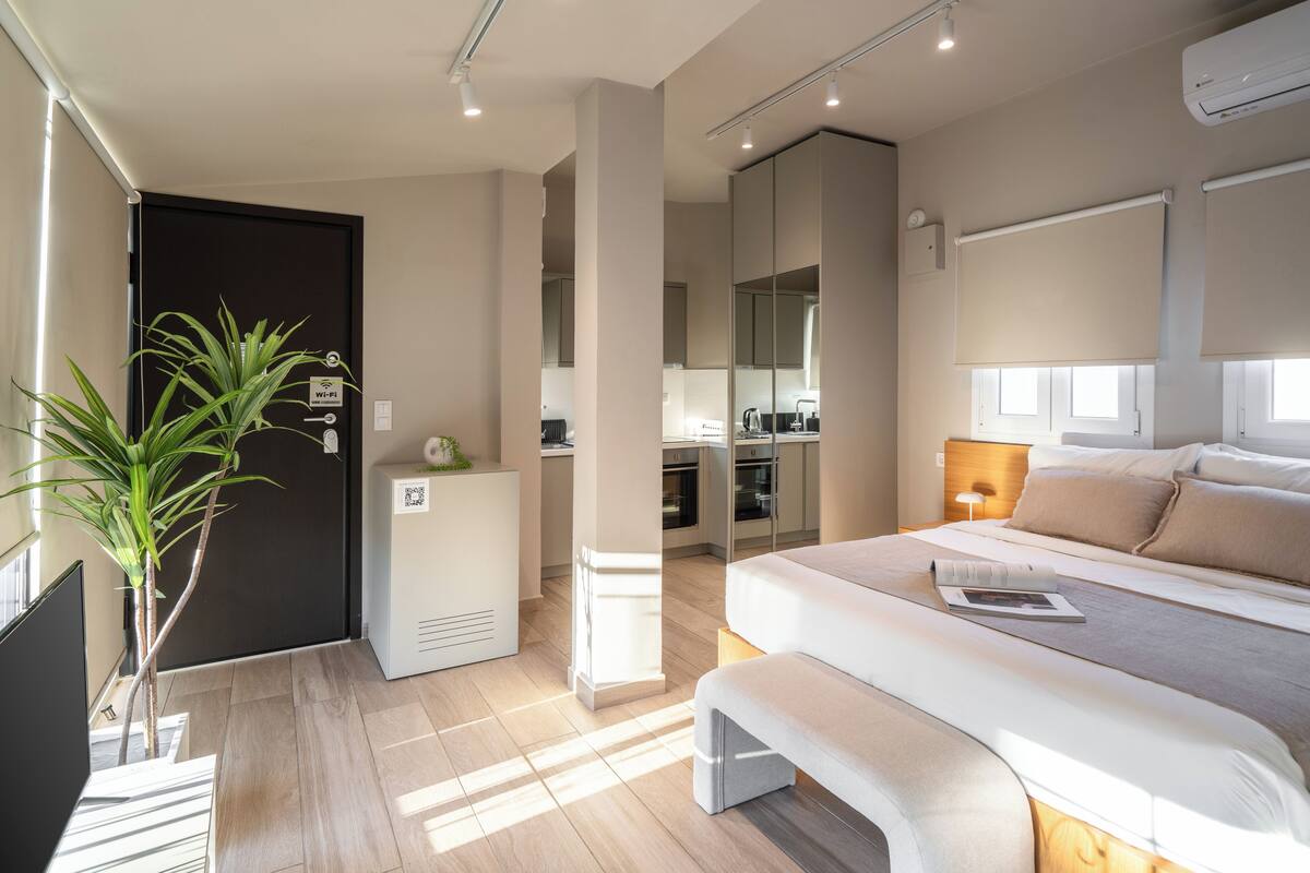Ein modernes Hotelzimmer mit einem großen Bett, einem Nachttisch und einer Zimmerpflanze. Im Hintergrund befindet sich eine Küchenzeile und ein separater Badebereich.