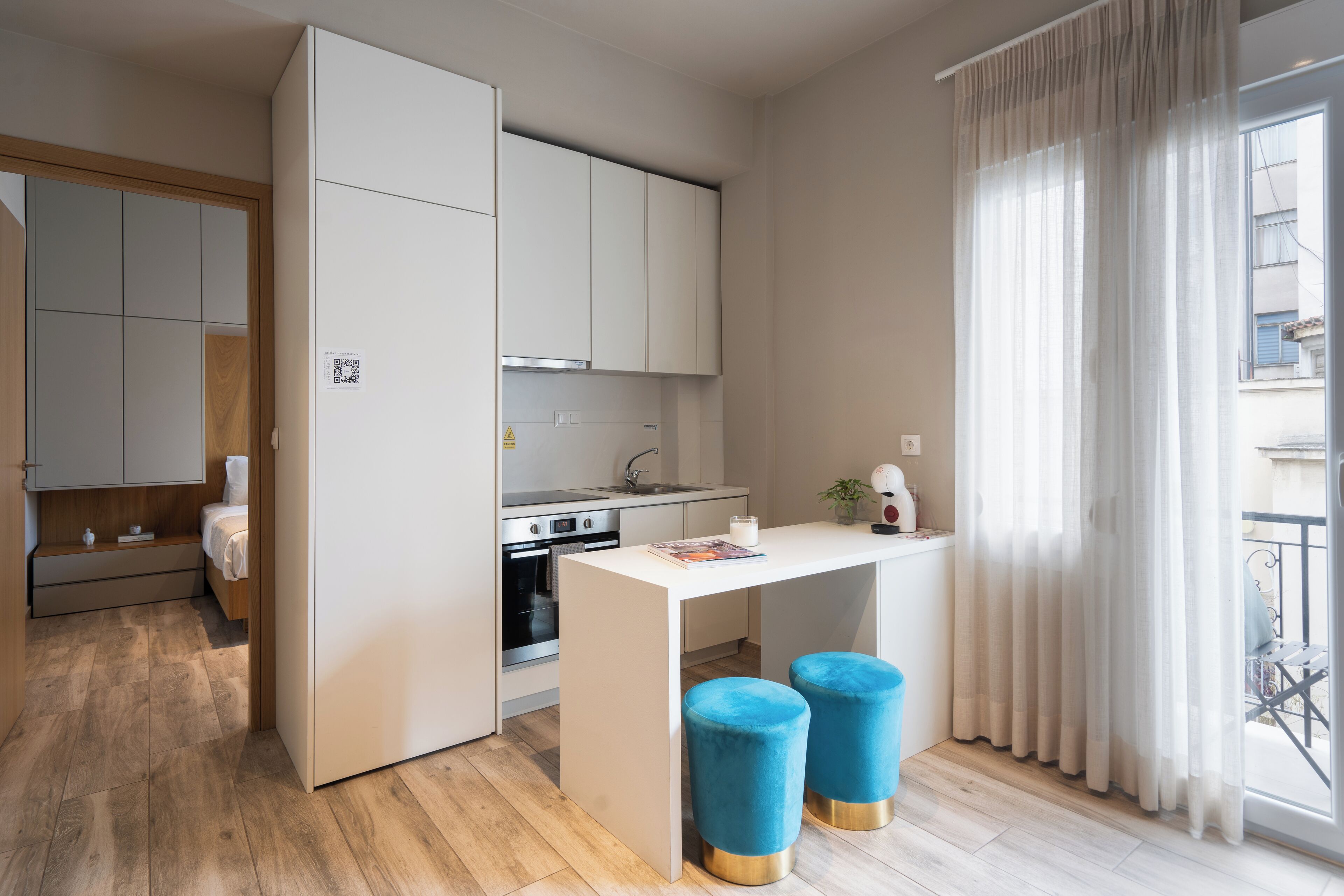 A compact kitchen with a central island, wooden flooring, and a view of the balcony through sheer curtains.