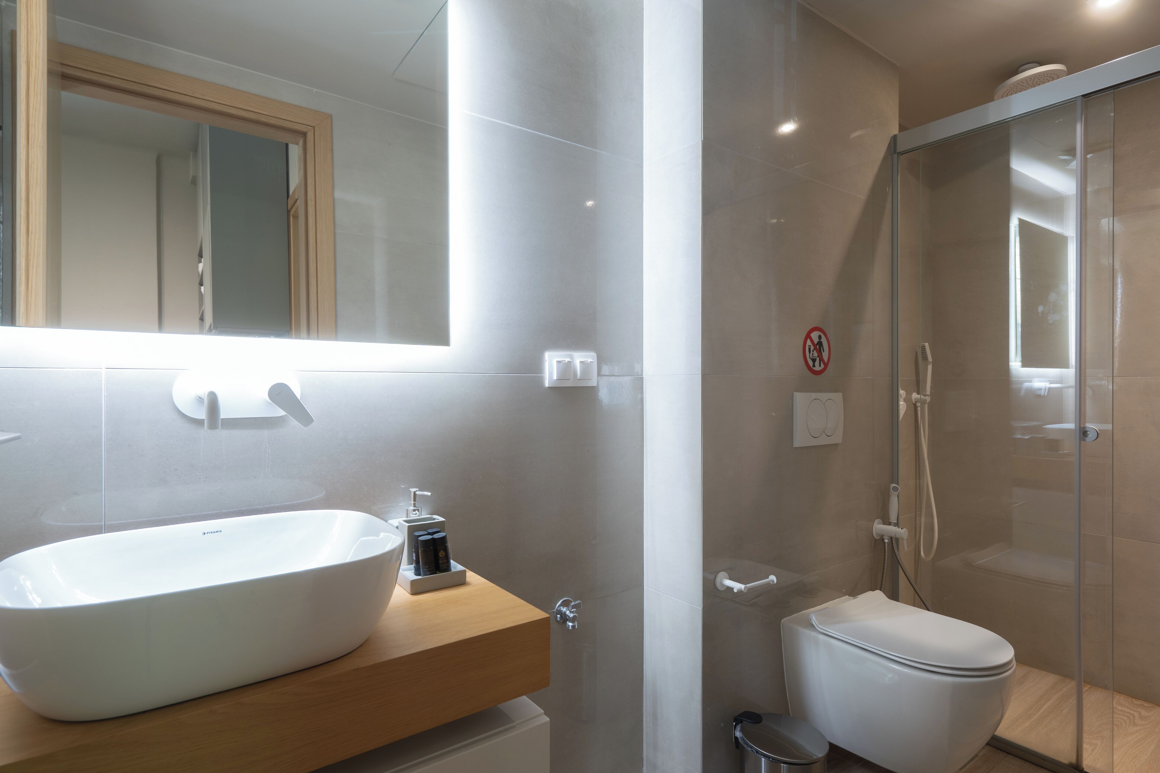 A modern bathroom with a large white vessel sink, a wooden vanity, a toilet, and a glass-enclosed shower area.