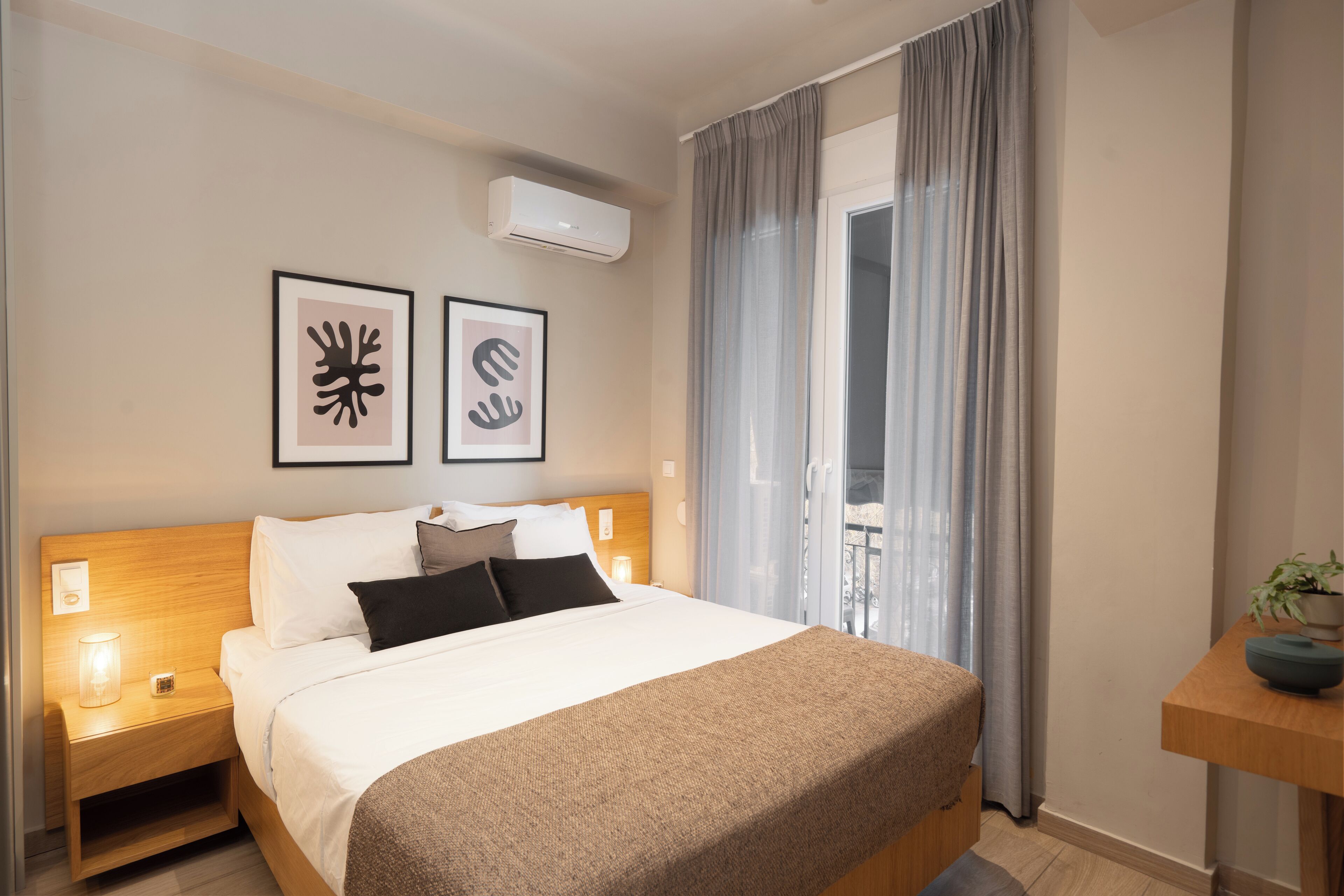 A hotel room with a bed, bedside tables, a wall-mounted air conditioner, and a view of a balcony through sheer curtains.
