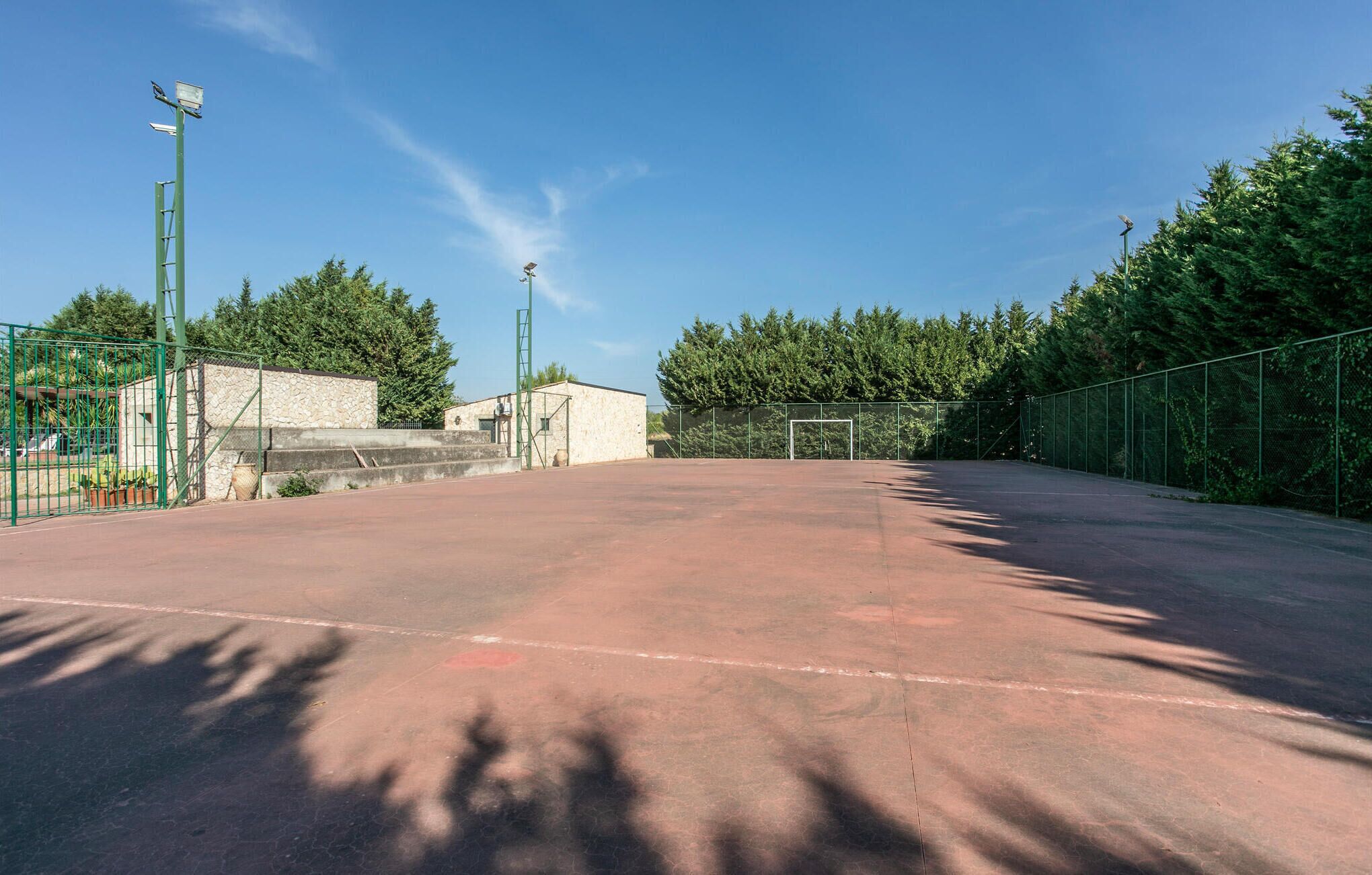 Tyhjä tenniskenttä, jossa on vihreä aita, kivirakennus ja taustalla puita.