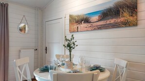 A dining area with a round table set for four, white chairs, a vase with flowers, and a painting of a beach scene on the wall. - Nice home in Heinkenszand with sauna (Heinkenszand)