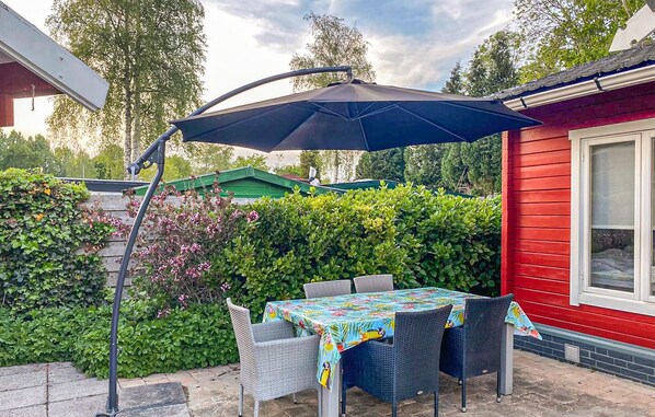 Een terras met een tafel en stoelen onder een grote parasol, omringd door groen en een rood gebouw.