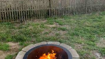 A circular fire pit with a fire burning in the center, surrounded by a grassy area and a wooden fence in the background.