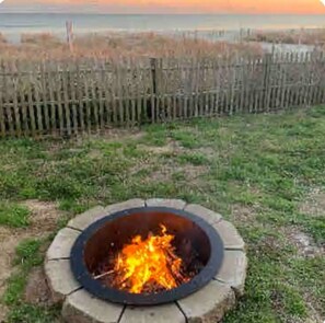 A circular fire pit with a fire burning in the center, surrounded by a grassy area and a wooden fence in the background.