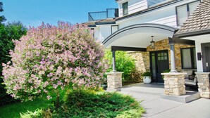 Une maison à deux étages, dotée d’une façade en pierre, d’un porche abrité et d’un jardin bien entretenu avec des arbres en fleurs.