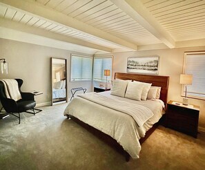 A bedroom with a large bed, a chair, a nightstand, a lamp, a mirror, and a window with blinds.