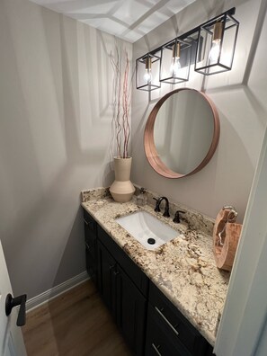 A bathroom with a white sink, dark cabinets, a marble countertop, a round mirror, and a vase with branches.
