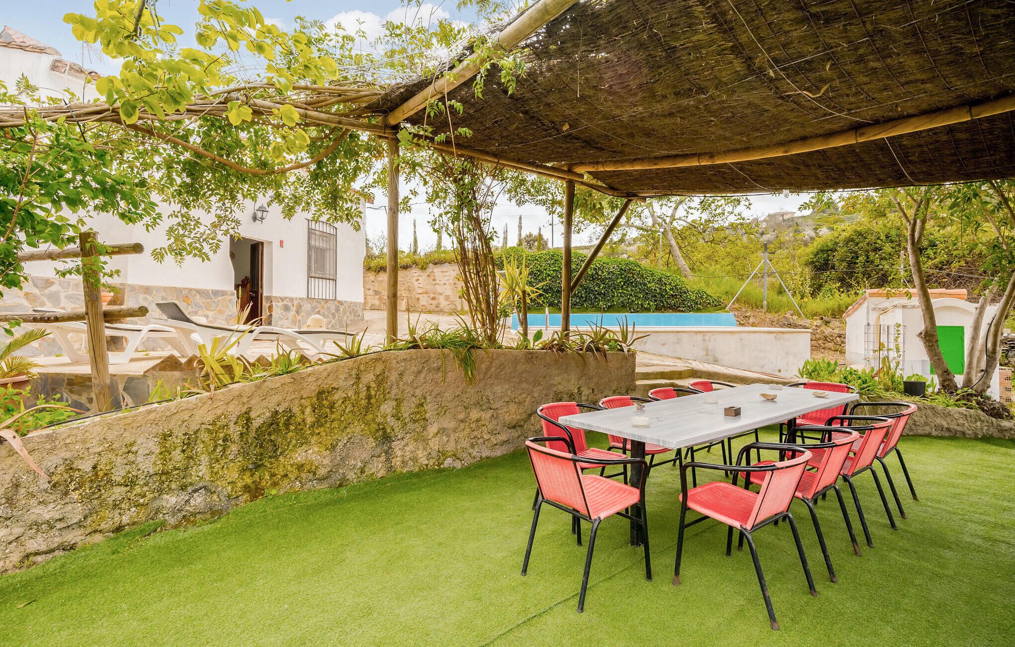 Un patio al aire libre con mesa y sillas, rodeado de follaje y un muro de piedra.