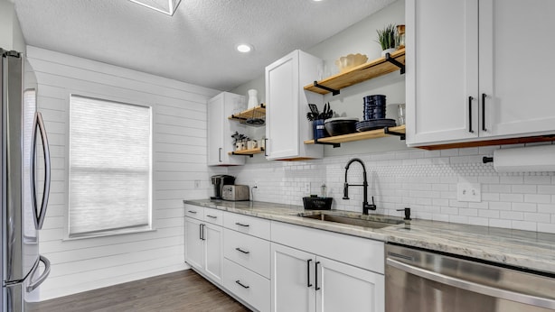 A modern kitchen with white cabinetry, a stainless steel refrigerator, a built-in dishwasher, and wooden shelves above the counter. - Pet Friendly Fenced Backyard Game Room Patio Firepit Trailer Motorcycle Parking (Myrtle Beach)