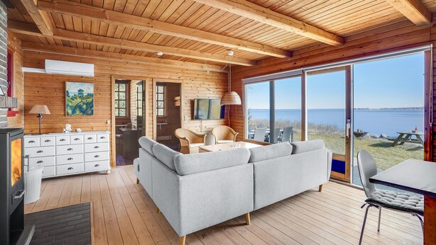 Ein gemütliches Cabin-Interieur mit Holzbalkendecke, einer Couch, einem Esstisch und Blick auf das Wasser.