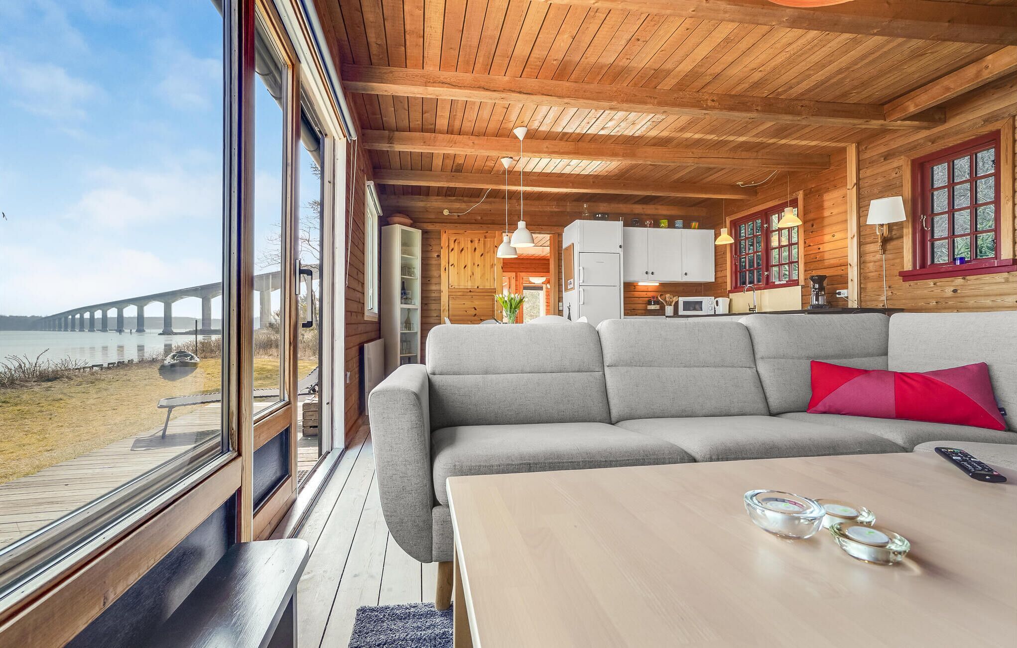 Une cabane dotée d’une grande fenêtre donnant sur un plan d’eau et un pont. À l’intérieur, on trouve un canapé gris, une table en bois et un coin cuisine avec des placards et un évier.