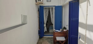 Un pasillo estrecho con una puerta azul que da a una habitación. Se ve una pequeña mesa con una silla y una planta en maceta dentro de la habitación.