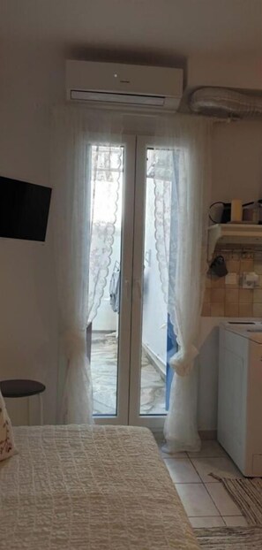 A room with a bed, a small table, an air conditioning unit, a washing machine, and a view through a glass door.