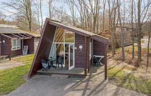 Una cabaña de madera con techo inclinado, puerta de cristal y un pequeño porche con mobiliario exterior.