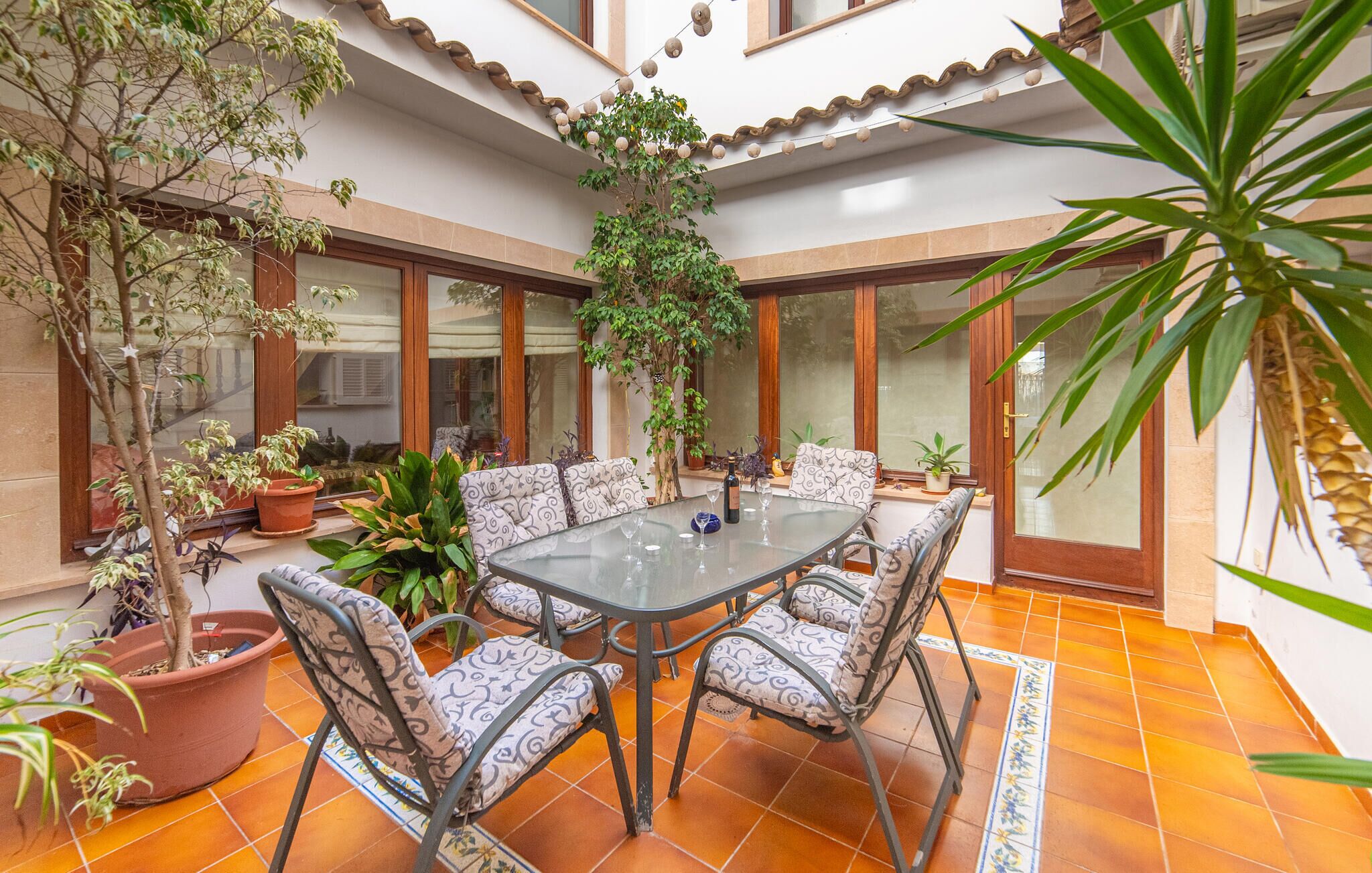 A spacious patio with a glass-top table surrounded by patterned chairs, potted plants, and a tiled floor.