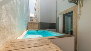 Una pequeña piscina rectangular con un deck de madera que la rodea, frente a un edificio de fachada blanca y una puerta de cristal.