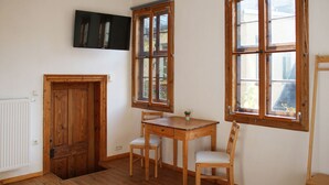 Una habitació amb una porta de fusta, una taula petita amb dues cadires i dues finestres amb marc de fusta.