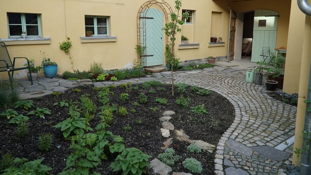 Un pati amb un camí pavimentat, una petita zona de jardí i un edifici amb una porta i finestres de blau clar.
