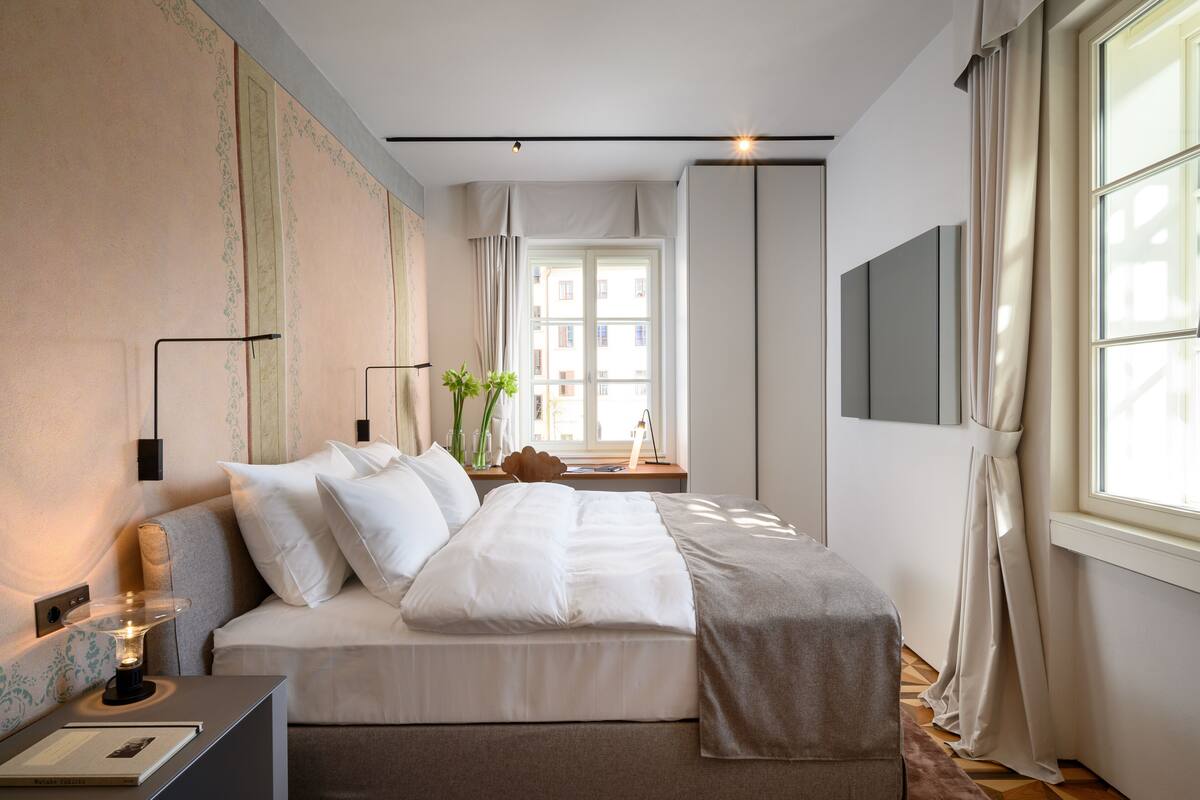 Ein modernes Hotelzimmer mit einem ordentlich bezogenen Bett, einem Nachttisch mit Lampe und einem großen Fenster mit Vorhängen.