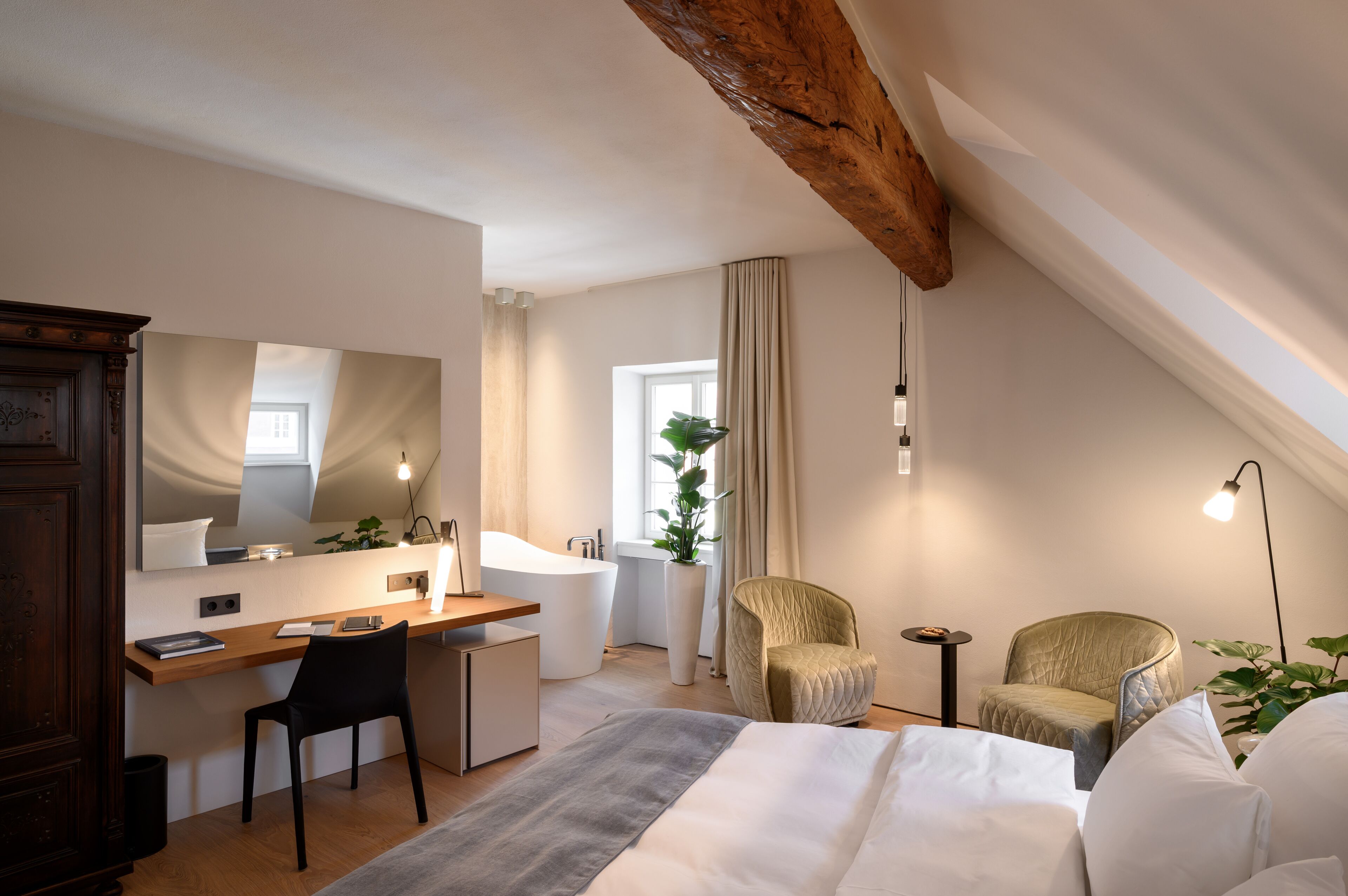 A modern bedroom with a wooden desk, a large bed, two chairs, a large mirror, and a plant.