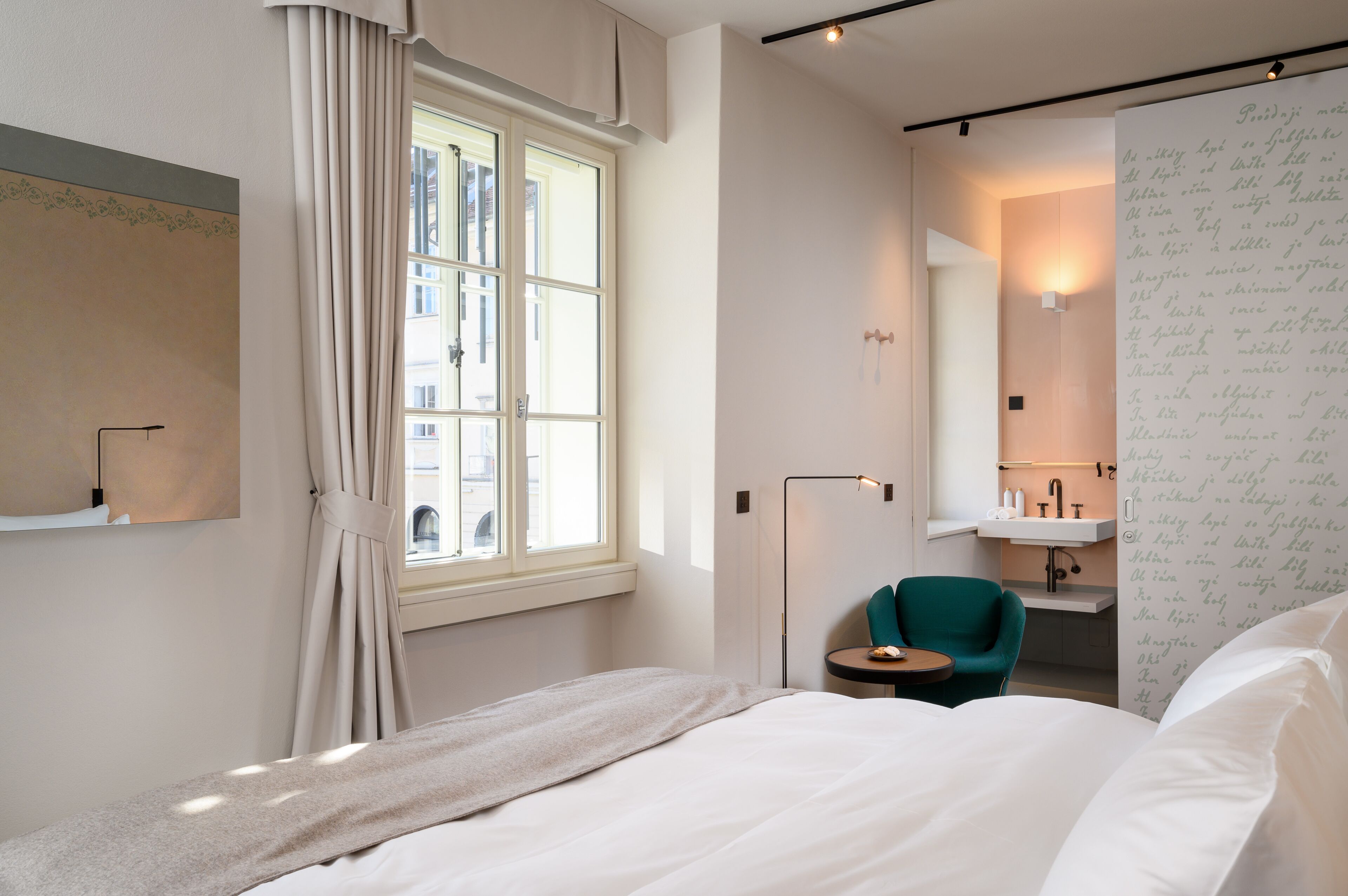 Quarto de hotel moderno com uma cama grande, uma mesa redonda pequena, uma cadeira verde-água, uma janela com cortinas, um espelho fixado na parede e um banheiro com pia e suporte para toalhas.