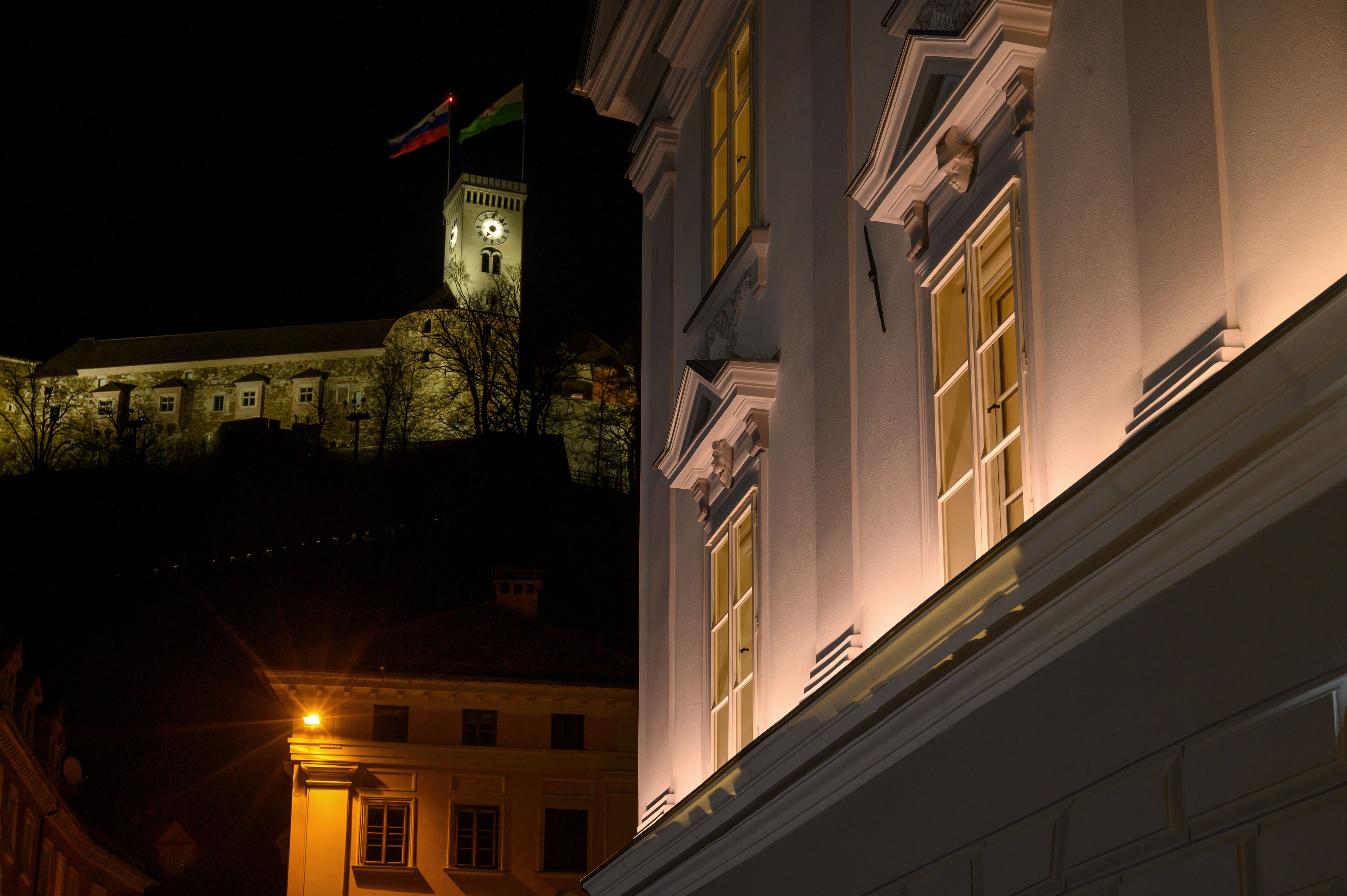 Un bâtiment doté d’une tour horloge et de drapeaux, présentant un style architectural classique avec des fenêtres disposées de manière symétrique et une façade bien éclairée.