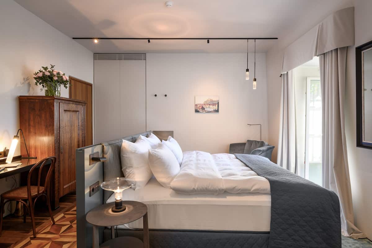 Ein modernes Hotelzimmer mit einem ordentlich bezogenen Bett, einem Schreibtisch mit Lampe, einem Sessel und einem kleinen Tisch ebenfalls mit Lampe.