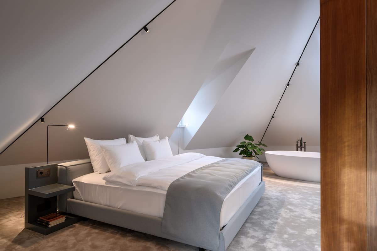 Ein modernes Schlafzimmer mit Schräge, einem großen Bett, einem Nachttisch, einer Leselampe, einer Pflanze und einer freistehenden Badewanne.