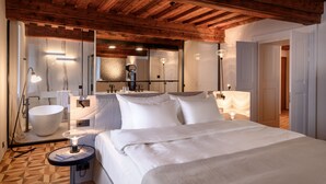 Quarto de hotel moderno com teto de madeira, uma cama grande, uma mesa de cabeceira redonda com abajur, uma banheira branca e um espelho grande.