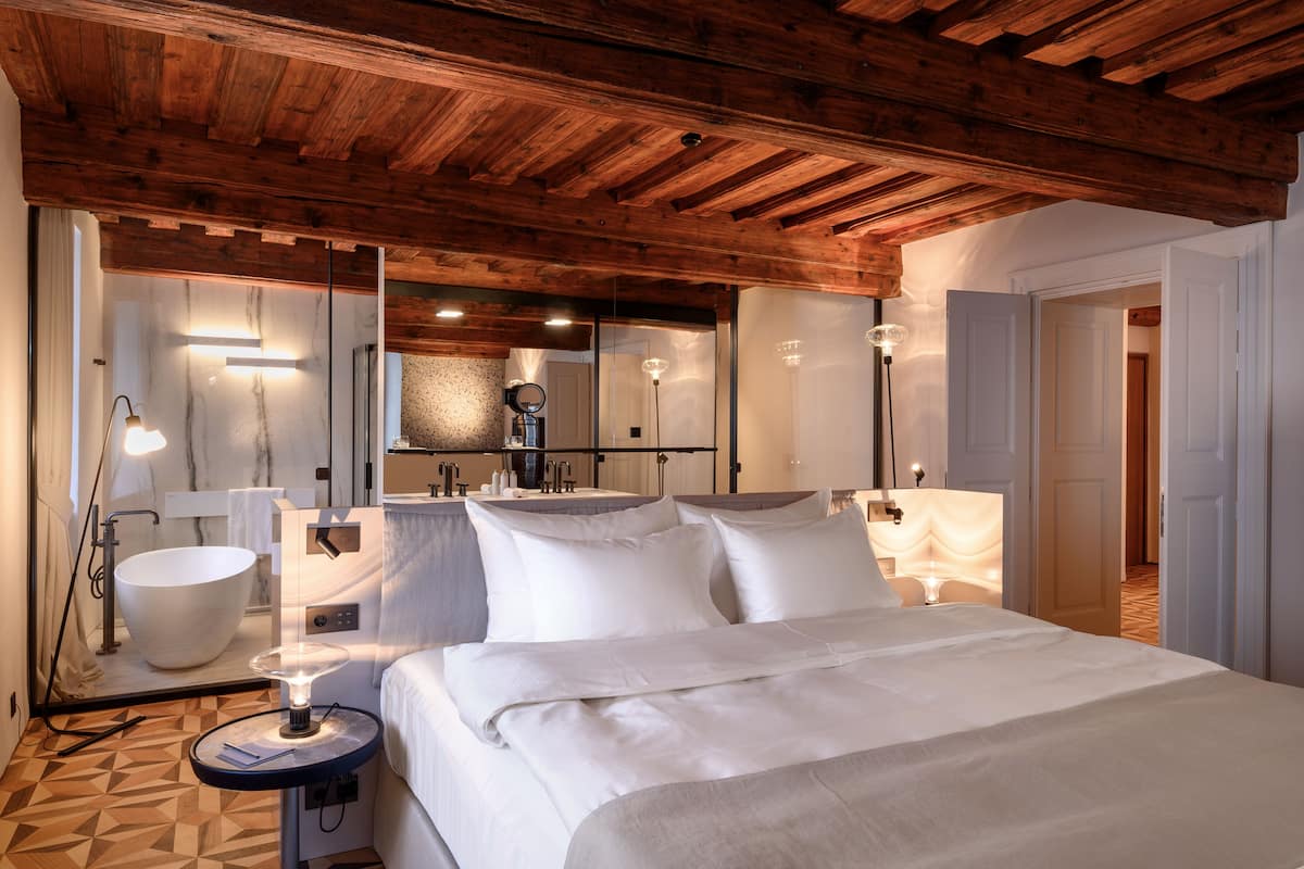 Ein modernes Hotelzimmer mit Holzboden, einem großen Bett, einem runden Nachttisch mit Lampe, einer weißen Badewanne und einem großen Spiegel.