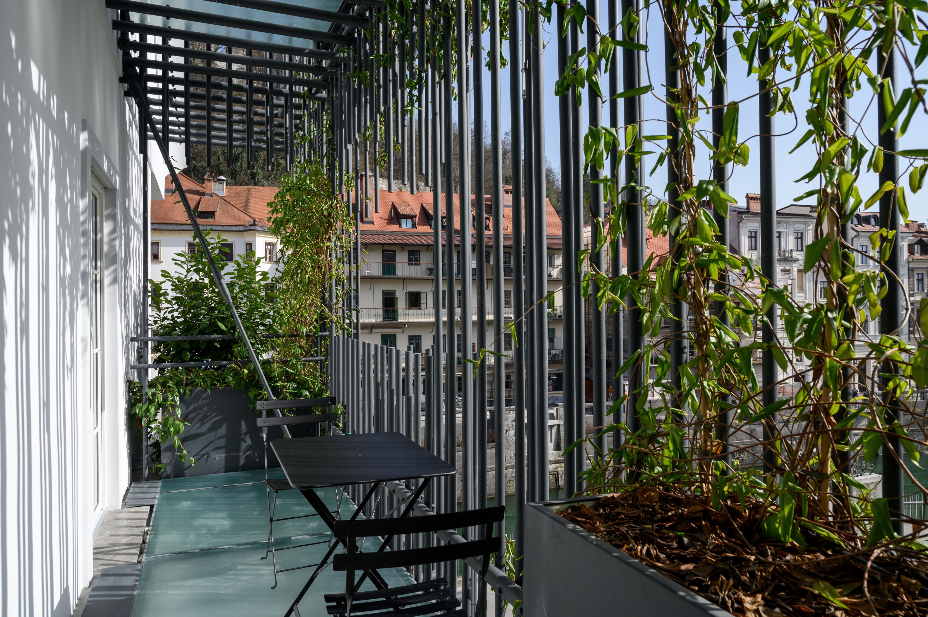 A balcony with a table and chairs, surrounded by vertical metal bars and greenery.