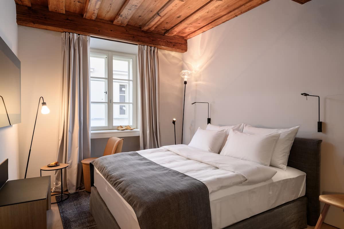 Ein Schlafzimmer mit Holzbalkendecke, einem Bett mit weißer Bettwäsche, einem Schreibtisch, einem Stuhl und einem Fenster mit Vorhängen.