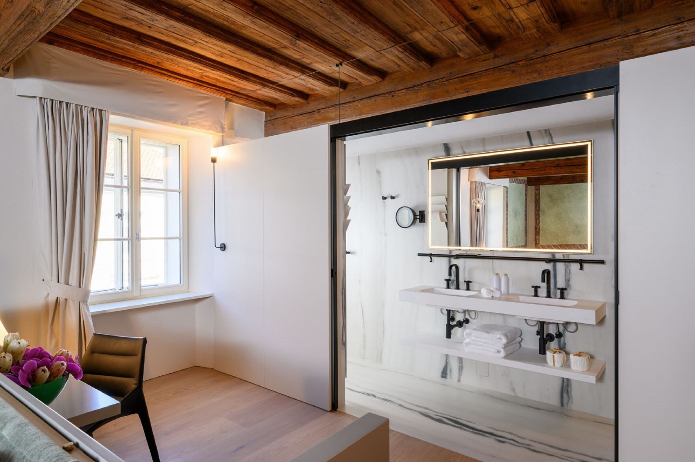 A modern bathroom with a large mirror, white marble sink, and wooden ceiling beams.