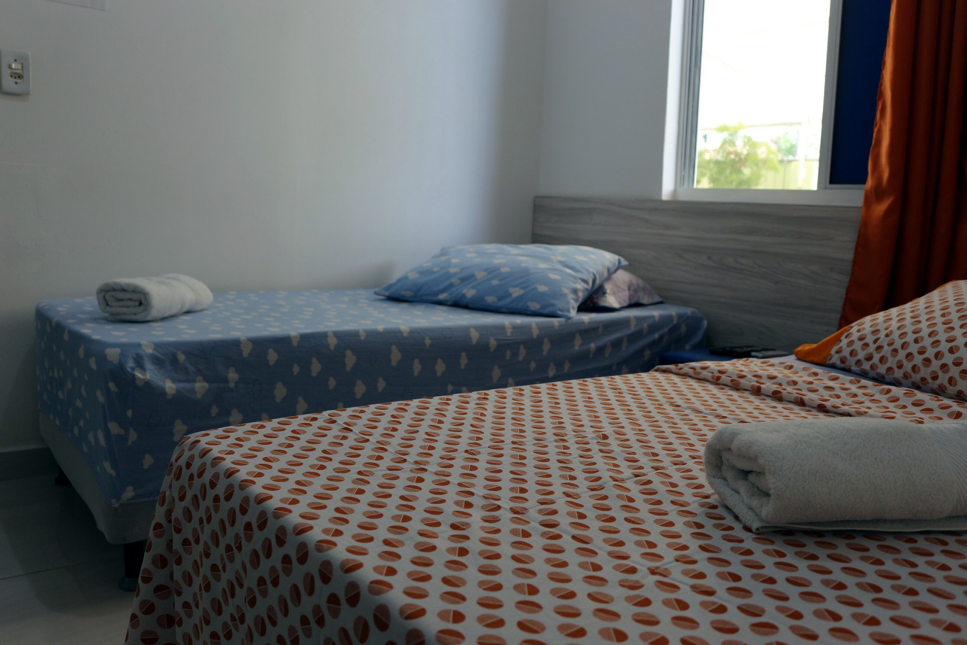 A hotel room with two beds, one with a blue bedspread and the other with an orange bedspread. There is a window with curtains, and a towel is placed on the bed with the blue bedspread.