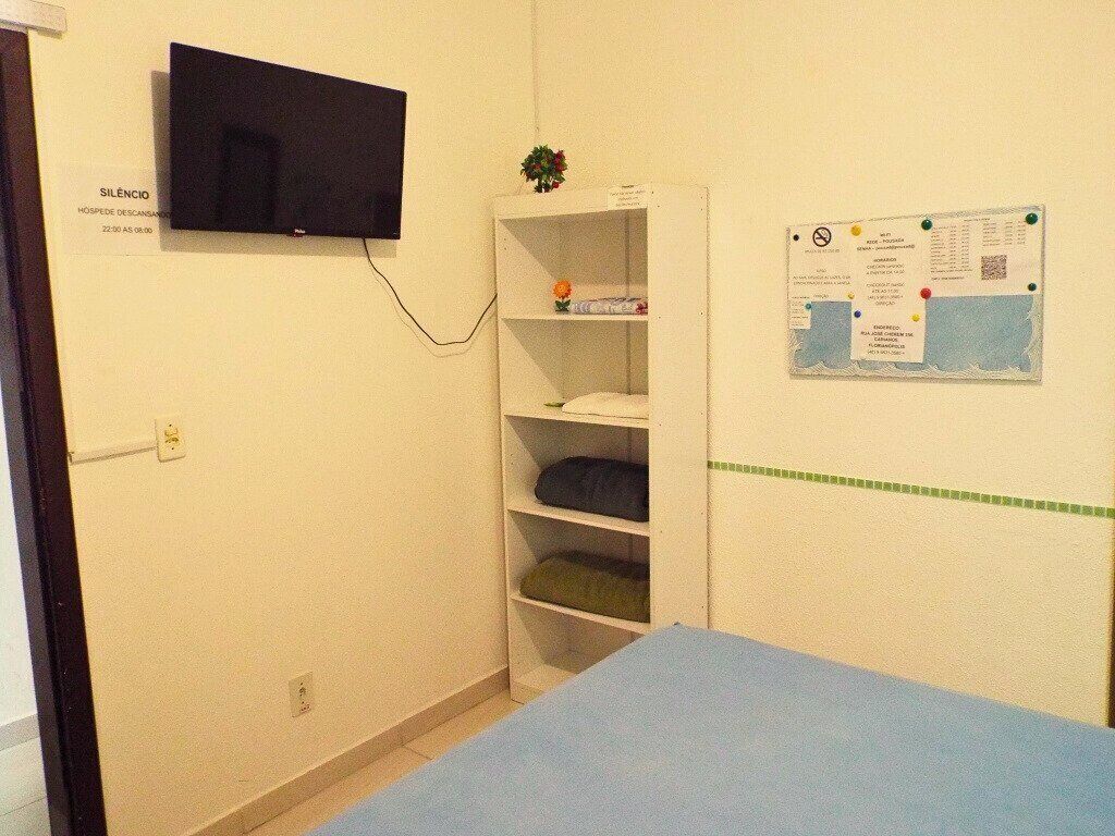 A room with a television mounted on the wall, a white shelving unit with folded items, a blue bed, and a bulletin board with various notices.
