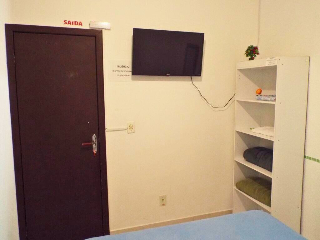 A room with a dark door labeled 'SAÍDA,' a flat-screen TV mounted on the wall, and a white shelving unit with folded items.