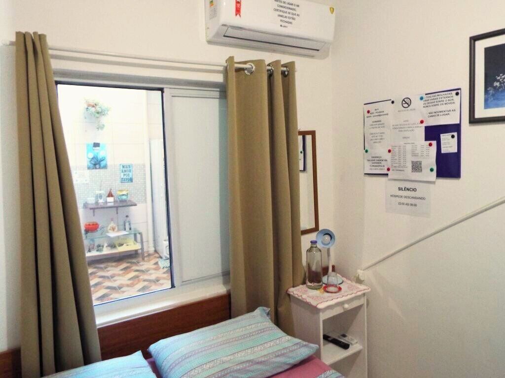 Quarto com cama, janela com cortinas, uma mesinha com garrafa e copo, e parede com alguns papéis.