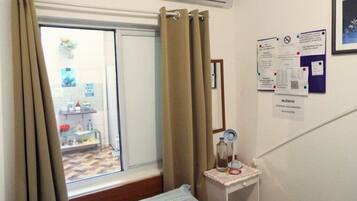 Quarto com cama, janela com cortinas, uma mesinha com garrafa e copo, e parede com alguns papéis.